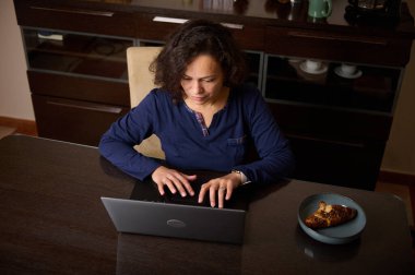 Koyu renk ahşap bir yemek masasında oturan bir kadın, modern bir mutfağın yakınındaki bir tabakta hamur işi otururken dizüstü bilgisayarla yazı yazıyor..