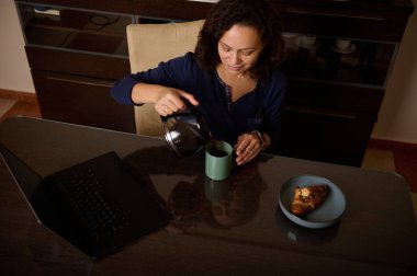 Ev ofisinden bir kadın, bir laptop açıkken ve kruvasan tabağa oturmuş, rahat ve verimli bir sabah rutini yakalarken naneli bardağa kahve dolduruyor..