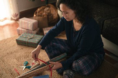 Ekose pijamalı bir kadın, hediyeleri paketlenmiş hediyeler ve ambalaj kağıtlarıyla sarar. Sıcak, şenlikli bir tatil hazırlığı sahnesi yaratır..