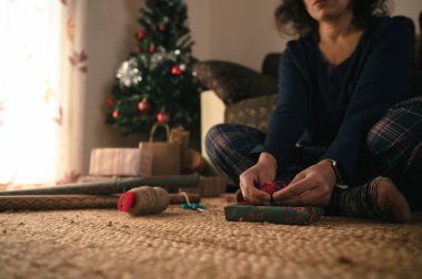Bir insan, hediyeleri renkli bir kağıt ve kurdeleyle süslenmiş bir Noel ağacının yanına sararak yerde oturuyor. Yumuşak doğal ışık ve sıcak, şenlikli bir atmosfer ev tatili hazırlıklarını iletir..