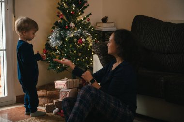 Bir anne, yanan bir Noel ağacının yanında yerde oturur ve sıcak bir tatil salonunda hediyeler arasında çocuğuna küçük bir hediye verir..