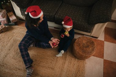 Bir anne ve çocuk Noel Baba şapkası takıyor, halının üzerinde oturuyor ve sıcak, şenlikli bir oturma odasında süsleri paylaşıyor..