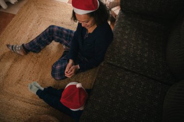 Oturma odasındaki halıya Noel Baba şapkası takmış bir ebeveyn ve çocuğun sıcak bir bayram anı, neşeli bir neşe, birliktelik ve rahat bir Noel atmosferi yakalıyor..