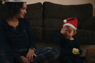 Bir anne ve çocuk Noel Baba şapkası takıyor, oturma odasında yerde oturuyor, evde bir lokma yemekle sakin, rahat bir Noel anını paylaşıyorlar..