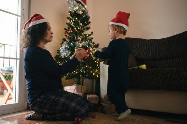 Noel Baba şapkası takan bir anne ve küçük bir çocuk sıcak bir oturma odasında Noel ağacı süsler. Altında hediyeler yatar ve yumuşak ışıklar neşeli, samimi bir tatil anı yaratır..