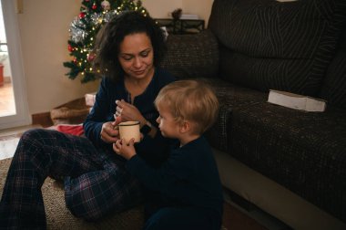 Bir anne ve küçük bir çocuk, bir Noel ağacının yanında yerde oturup, rahat bir oturma odasında bir kupayı paylaşıyorlar..