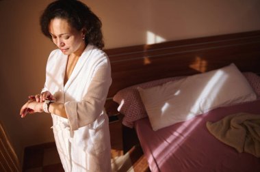 Güneş doğarken beyaz bornozlu bir kadın bir yatağın yanında duruyor, saatini kontrol ediyor. Ilık sabah ışığı günlük rutin ve kişisel bakım için sakin ve samimi bir sahne yaratır..