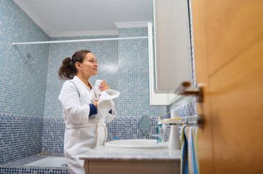 Beyaz bornozlu bir kadın lavabonun yanında havlu tutuyor, mavi mozaikli bir banyonun önünde dikiliyor..