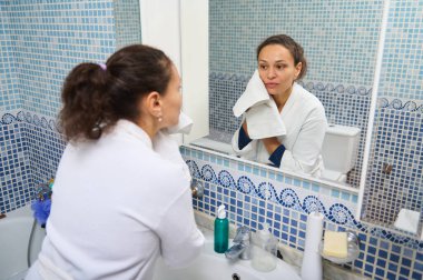 Beyaz bornozlu bir kadın elinde havluyla mavi mozaik banyo aynasında yansımasına bakıyor. Sahne kişisel bakım, rutin ve sessiz yansıma içeriyor..