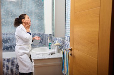 Beyaz bornoz giyen bir kadın banyo lavabosuna losyon sürüyor. Parlak, fayanslı bir banyoda cilt bakımlı bir güne hazırlanıyor..