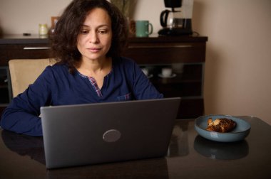 Düşünceli bir kadın yemek masasında dizüstü bilgisayarına odaklanmış oturuyor. Yakınlarda bir tabakta kruvasan olması sıradan bir ev işi anı olduğunu gösterir..