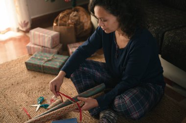 Ekose pijamalı bir kadın, hediyeleri paketlenmiş hediyeler ve ambalaj kağıtlarıyla sarar. Sıcak, şenlikli bir tatil hazırlığı sahnesi yaratır..