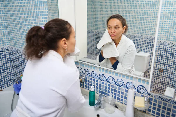 Beyaz bornozlu bir kadın elinde havluyla mavi mozaik banyo aynasında yansımasına bakıyor. Sahne kişisel bakım, rutin ve sessiz yansıma içeriyor..