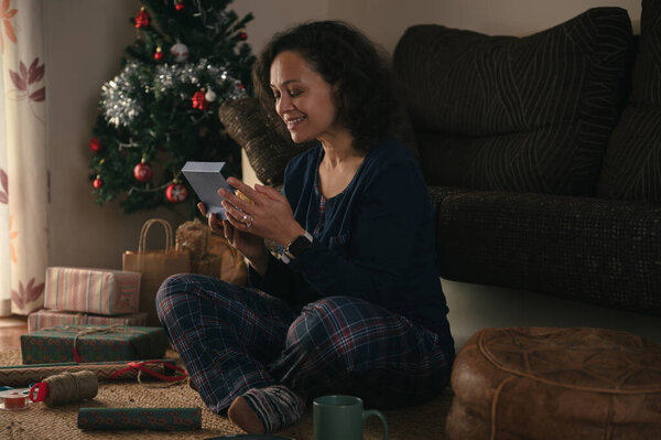 A warm, homey holiday moment: a woman on the rug nearby a decorated Christmas tree, inspecting a small gift, with wrapped presents and festive decor around.