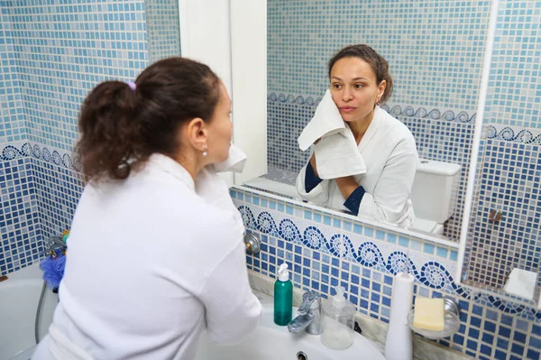 Beyaz bornozlu bir kadın elinde havluyla mavi mozaik banyo aynasında yansımasına bakıyor. Sahne kişisel bakım, rutin ve sessiz yansıma içeriyor..