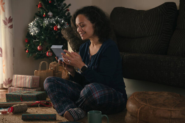 A warm, homey holiday moment: a woman on the rug nearby a decorated Christmas tree, inspecting a small gift, with wrapped presents and festive decor around.