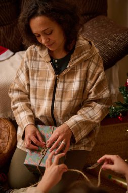 Ekose ceketli bir kadın, hediyesini sicimle sarıp sıcak bir oturma odasında aile hediyesini paylaşır..