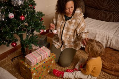 Bir anne süslü bir Noel ağacının yanında diz çöker, süslere ve hediyelere hayran olan bir çocuğa rehberlik eder. Sıcak, samimi aile sahnesi, sıcak ve şenlikli tatiller..