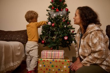 Evdeki Noel ağacını süsleyen anne ve çocuğun sıcak, samimi bir sahnesi. Altında hediyeler oturuyor, ışıklar yanıp sönüyor, ve ikisi de neşe, bağ ve tatil ruhunu paylaşıyor..
