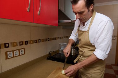 Bej önlüklü bir adam, temiz bir mutfakta, ahşap bir tahtanın üzerinde soğan doğruyor ve akşam yemeğini hazırlamaya odaklanıyor..