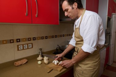 Odaklanmış bej önlüklü bir adam mutfak tezgahında mantar doğruyor. Sahne ev yemeklerini, aile yemeklerini ve günlük aşçılık çabalarını anlatıyor..