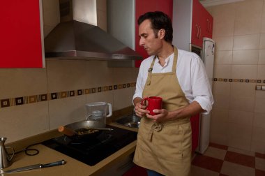 Çağdaş bir mutfakta yemek pişirirken elinde kırmızı bardakla bej önlüklü bir adam sobanın yanında duruyor. Sıcak, aile içi olaylar ev hayatını ve aile yemeklerini akla getiriyor..