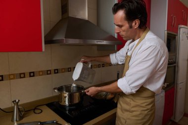 Önlüklü bir adam ocakta yemek pişirir, çorba yapmak için tencereye su döker. Kırmızı dolaplı temiz mutfak sıcak, ev yapımı bir an yaşatıyor..