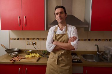 Bej önlüklü bir adam kırmızı bir mutfakta kollarını kavuşturup yemek yapmaya hazır bekliyor. Tezgahtaki patates ve kaseler ev yemeklerini ve aile yemeklerini akla getiriyor..