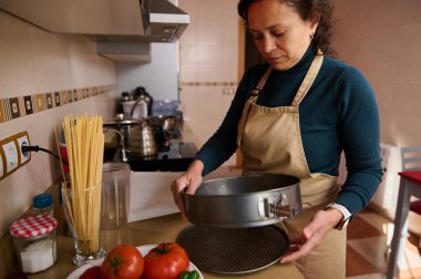 Önlüklü bir kadın mutfak tezgahında elinde yuvarlak bir tavayla duruyor. Taze domatesler, makarna, kavanozlar ve çatal bıçaklar ev yemeği hazırlarken onun etrafını sarar..