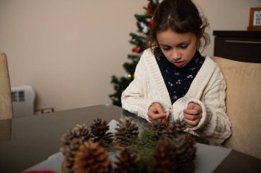 Genç bir kız Noel için çam kozalağı süslemeleri yapıyor. Arka planda Noel ağacı olan sıcak bir ev sahnesi, sıcak aile tatili anları yakalıyor..