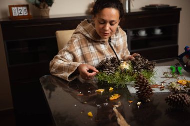 Ekose ceketli bir kadın mutfak masasında yeşil çelenklerle çam çelengi şekillendiriyor. Sıcak, şenlikli bir ev işi anı yaratıyor..