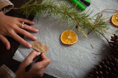 Sıcak bir tatil sahnesinde, çam iğneleri, kurutulmuş portakal dilimleri ve kozalaklarla çevrili kahverengi kağıda el yazısıyla yazılmış bir Noel etiketi yazılıyor..