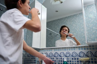 Genç bir çocuk banyo aynasında dişlerini fırçalıyor. Arka planda mavi mozaik fayanslar var. Her gün sakin bir anı yakalıyor..