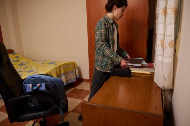 Genç çocuk, rahat bir yatak odasındaki ahşap bir masada, yanında bir sırt çantası ve bir yatakla, sessiz bir çalışma anını yakalayan defterler düzenliyor..