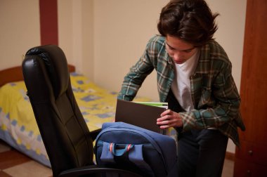 Genç bir çocuk, içinde not defterleri ve klasörler olan sırt çantasını rahat bir yatak odasına yerleştirir ve okula hazırlanır. Sahne rutin, çalışma, gençlik ve hazırlık içeriyor..