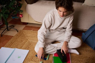 Odaklanmış genç bir çocuk, örülmüş bir halıya oturmuş, yakınlardaki bir Noel ağacıyla sıcak bir evde defter ve kalem kutusu düzenliyor..
