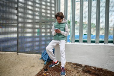 Genç bir çocuk metal bir çitin yanında oturuyor. Sırt çantasıyla telefonuna mesaj atıyor. Yeşil bir yelek ve günlük kıyafetler giyiyor, spor salonunda mola veriyor..