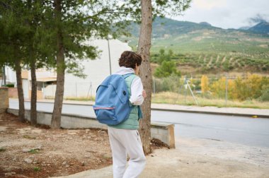 Mavi sırt çantalı genç bir çocuk, arka planda tepeler ve dağlarla birlikte, ağaçlarla kaplı bir yolda yürüyor, keşif ve gençlik taşıyor..