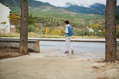 Mavi sırt çantalı genç bir çocuk ağaçların yanında sessiz bir yolda yürüyor, arka planda kayan tepeler ve dağlar var, gençlik ve açık hava keşiflerini yakalıyor..