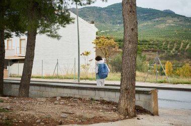 Mavi sırt çantalı genç bir çocuk arka planda zeytin tepeleri olan beyaz bir binanın yanında sessiz bir yol boyunca yürüyor..