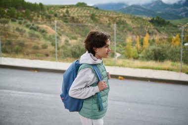 Mavi sırt çantası ve yeşil yeleği olan genç bir çocuk çitli bir yolda yürüyor, gündelik bir açık hava gününün tadını çıkarıyor..