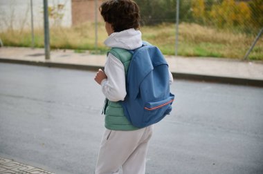 Mavi sırt çantalı genç bir çocuk okuldan sonra şehir caddesinde kapüşonlu ve yelekli yürüyor. Gençlik, gündelik ve günlük şehir hayatı..