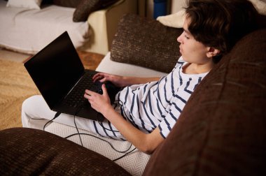 Genç bir çocuk kanepede rahatlar, rahat bir oturma odasında dizüstü bilgisayarda oyun oynar, gündelik gençlik hayatını yakalar ve oyun oynar..