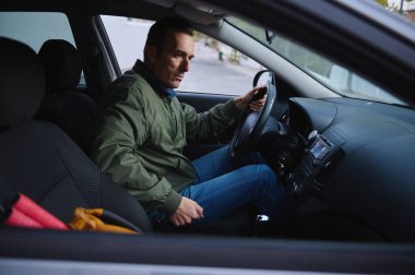 Sakin bir adam, yeşil ceket ve kot pantolon giymiş, modern bir arabanın direksiyonunda oturur. Yollara odaklanıyor, günlük, günlük bir sürüş anını yakalıyor..