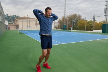 Mavi bluzlu formda bir adam ve lacivert şortlu bir adam antrenmana hazır bir tenis kortunda elleri başının arkasında geriniyor. Çitli ve ağaçlı açık hava manzarası.