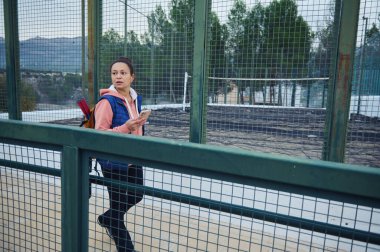 Sırt çantalı sıradan bir kadın yeşil bir çit boyunca yürüyor, bir elinde akıllı bir telefon, ağaçların arasında ve uzak tepelerin arasında açık hava parkında ya da spor alanında..