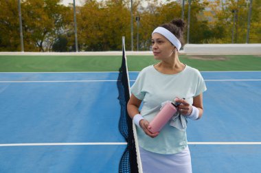 Saç bandı takmış odaklanmış bir kadın antrenmandan sonra elinde pembe bir su şişesi ve havluyla mavi bir tenis kortunda duruyor. Sonbahar ağaçları ve çitler açık hava sporları sahnesi yaratıyor..