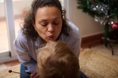 Bir kadın, arka planda Noel ağacı olan rahat bir oturma odasında bir çocuğu alnından nazikçe öper. İçtenlik, şefkat ve bayram aile sevincini aktarır..
