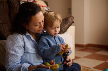 Sevgi dolu bir anne bebeğiyle oturur, yumuşak mavi tonlarda, bir aperatifi paylaşır ve muzları keşfederler. Aile bağlarını, bakımlarını ve günlük birlikteliklerini aktaran sıcak, samimi bir ev sahnesi..