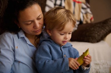 Sevgi dolu bir anne evde çocuğuyla oturur ve bir muza odaklanır. Sıcak ışık, samimi an, günlük aile hayatı, bağlanma ve bakım.